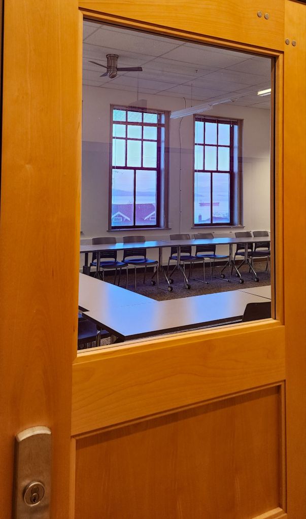 a photo of a classroom door. There is a window in the door. Behind the window are tables and chairs set up in a square and behind that are two more windows, through which is a view of the Columbia river.