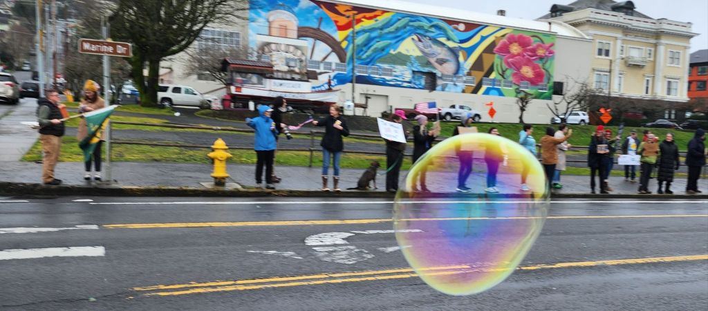 A color photo of a sparse crowd of protestors in front of a colorful mural. In the foreground is a huge soap bubble.