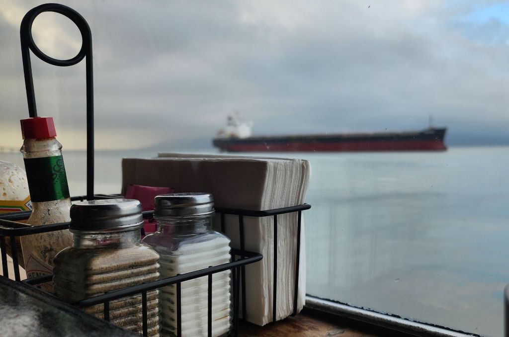 a photo of salt and pepper shakers and napkins; through the window behind them a huge cargo ship is proceeding along the river