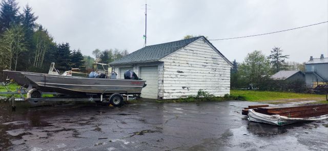 a rainy landscape featuring a boat shed with peeling paint, a boat on a trailer, some lumber in the driveway in the rain.