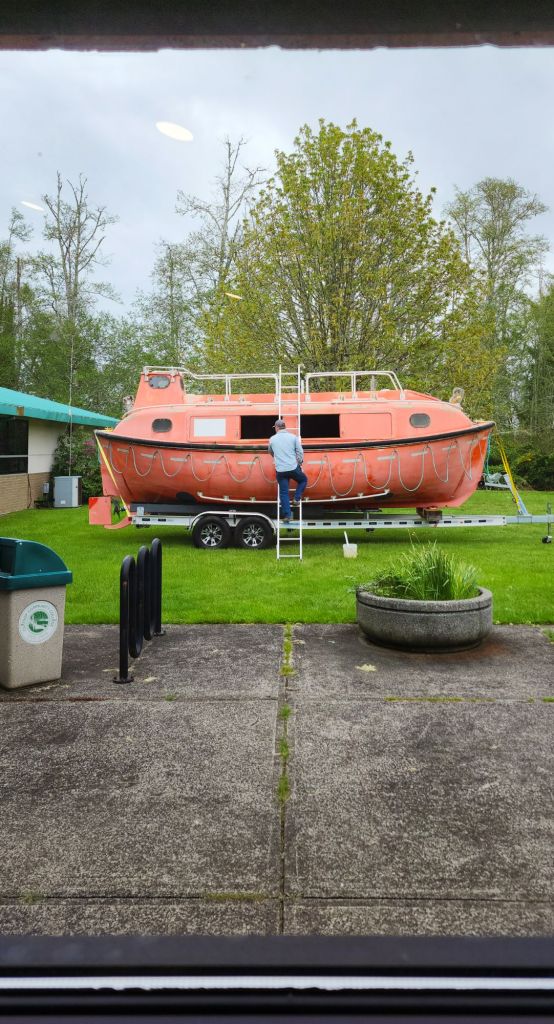 A man climbs a ladder leaning on an orange lifeboat on a trailer in the grass