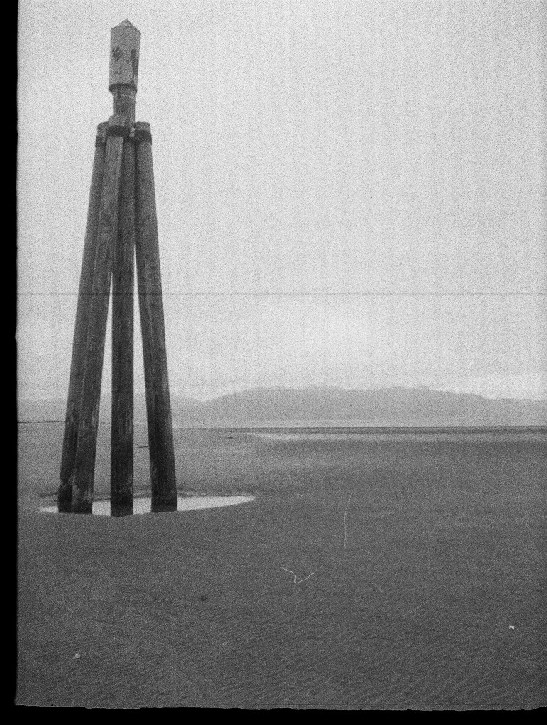 a black and white photo of a huge tripod holding up a buoy; it is low tide and the whole thing is visible