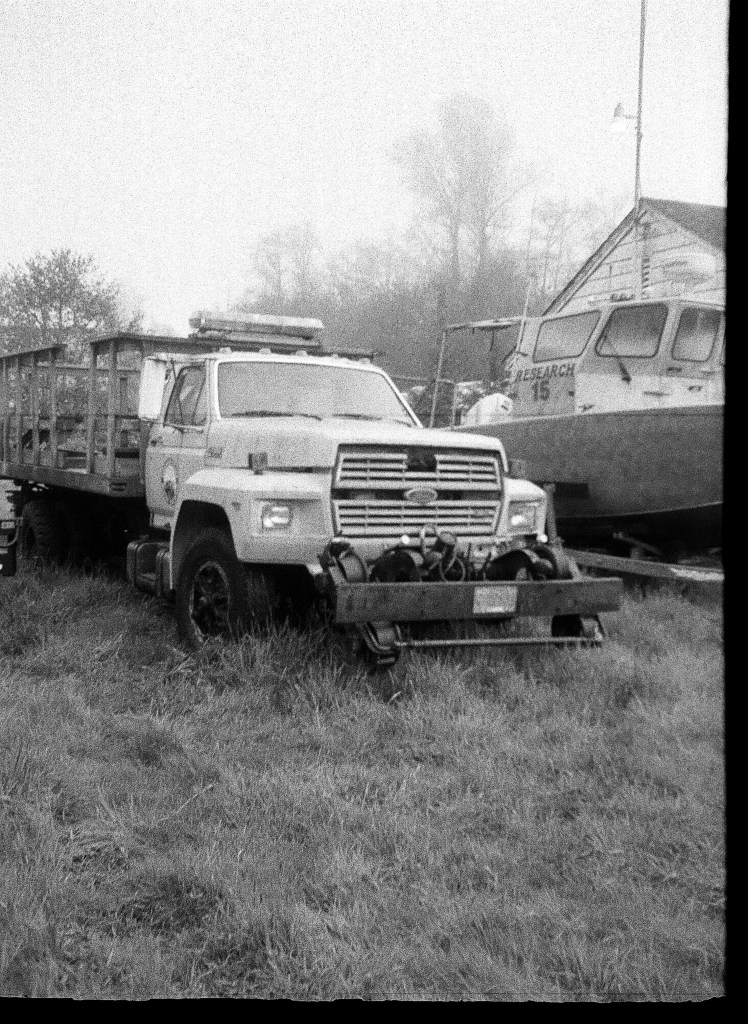 a black and white photo of a truck and a boat on a trailer, parked in the grass on a misty day. They look like they've been there a long time