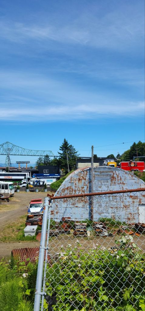 a view of a bit of Astoria including a chainlink fence, a rusted quonset hut, some old cars and in the background the Megler Bridge